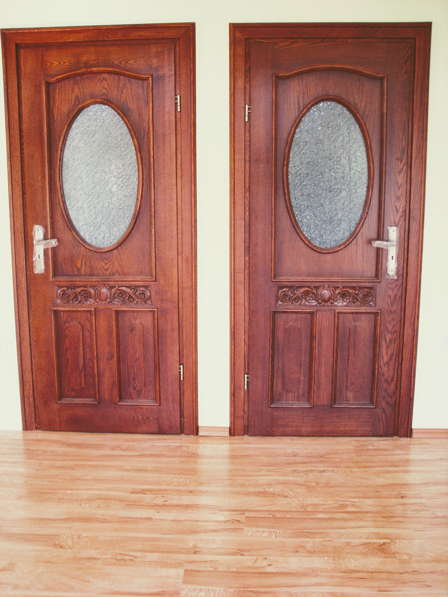Dwie drewniane, symetryczne drzwi wewnętrzne z owalnymi szybami z ornamentami, zamontowane obok siebie na tle jasnej ściany i paneli podłogowych.