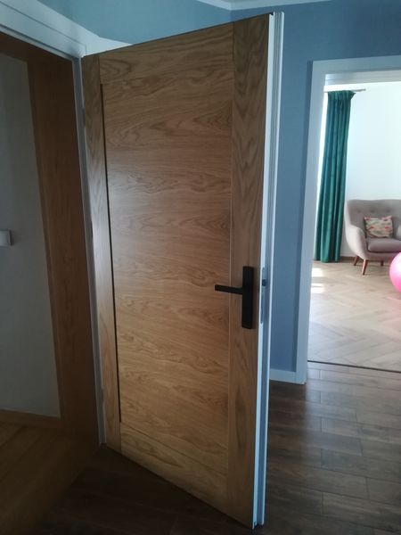 Otwarta, dębowa płycinowa drzwi wewnętrzne z czarną klamką, widok na fragment pokoju z szarą sofą i turkusowymi zasłonami.