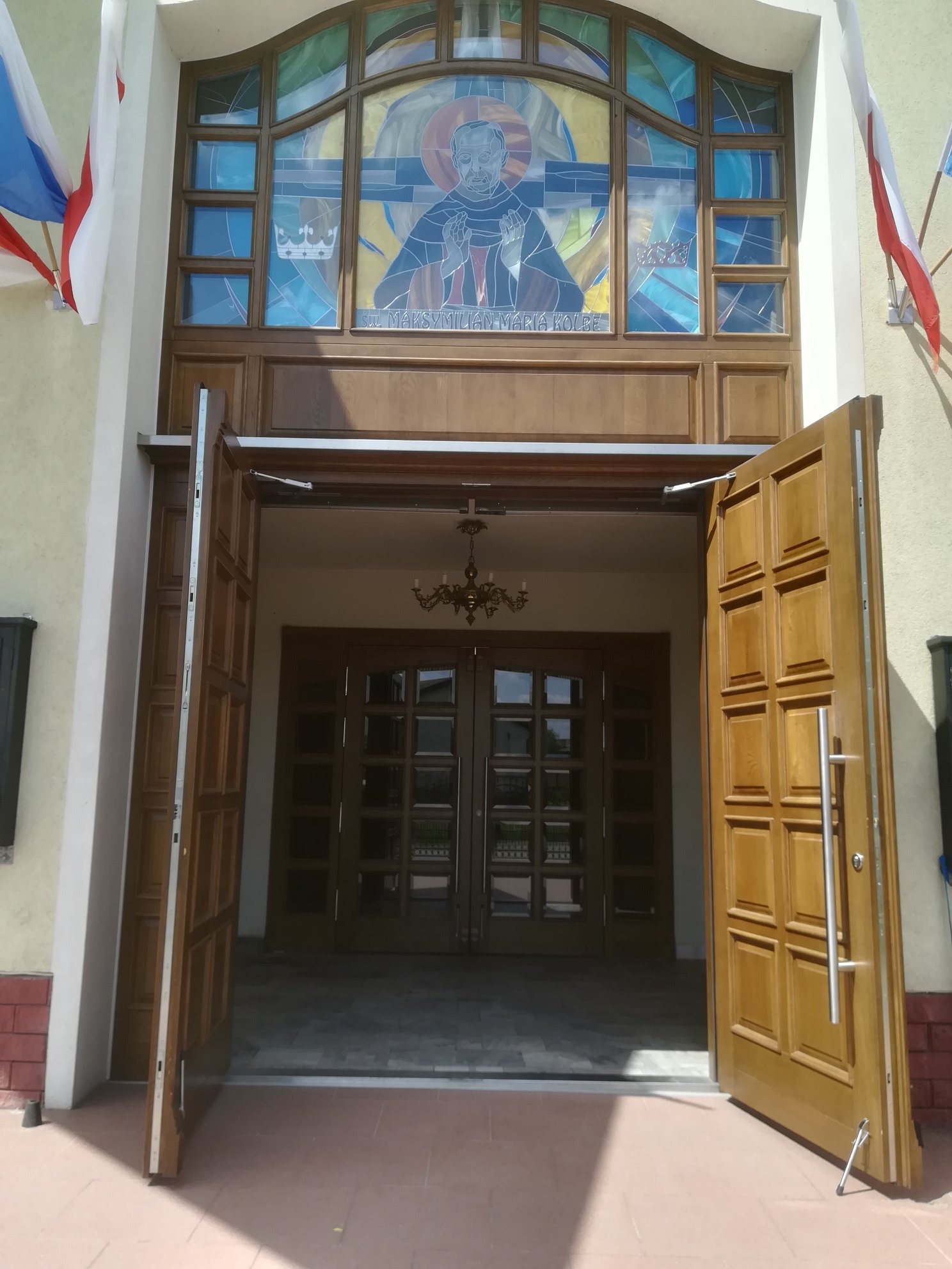 Wejście do budynku z otwartymi, drewnianymi drzwiami i witrażem przedstawiającym postać świętego nad nimi, zwieńczone flagami.