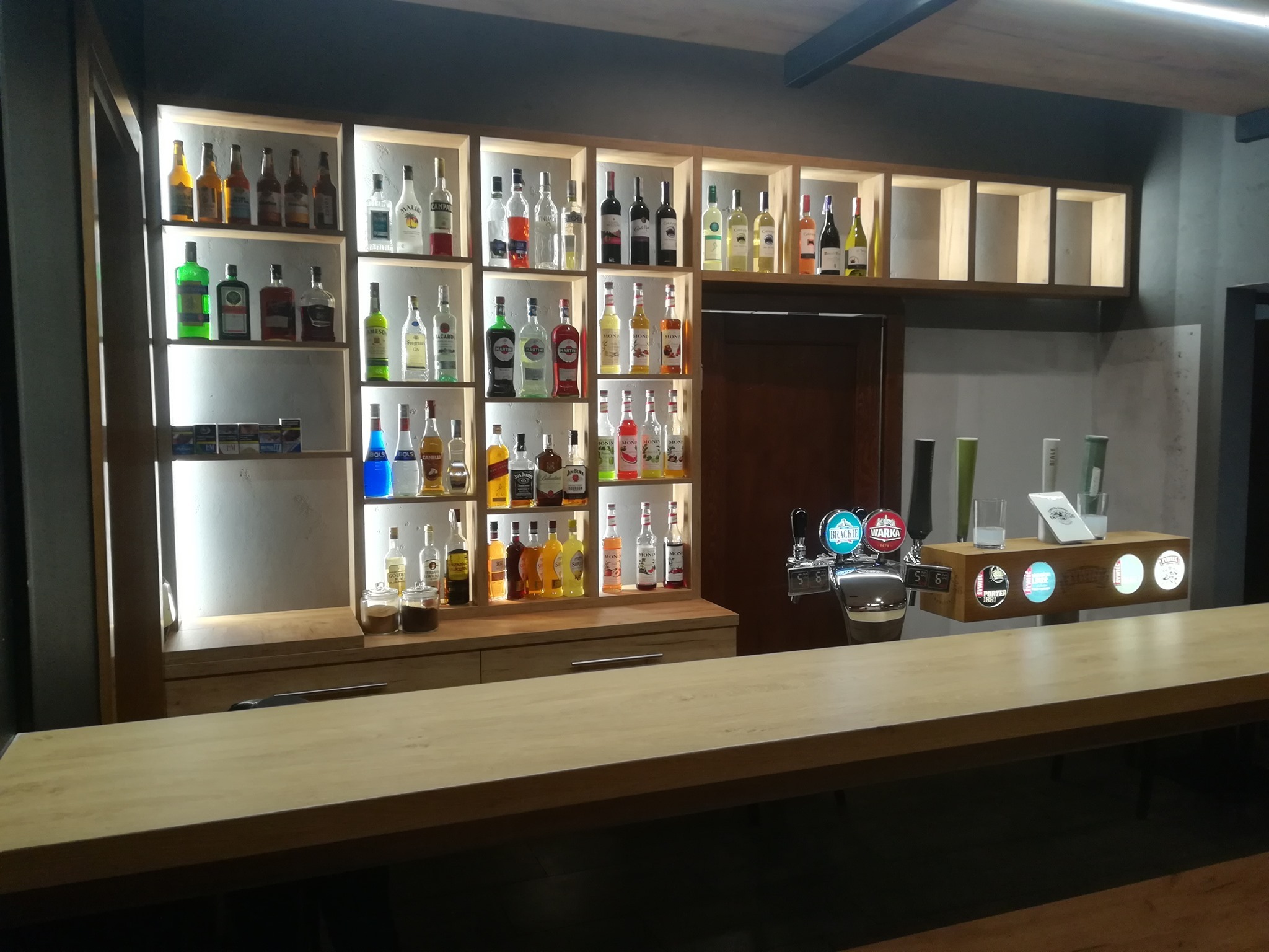 Drewniane regały z podświetleniem, wypełnione butelkami alkoholi, nad blatem baru w Jawiszowicach. Widoczne dystrybutory piwa.