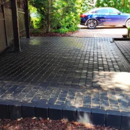 Ułożona kostka brukowa w kolorze grafitowym tworząca podjazd, z widocznym odwodnieniem liniowym i fragmentem drewnianej konstrukcji zadaszenia, na drugim planie zaparkowany niebieski samochód.