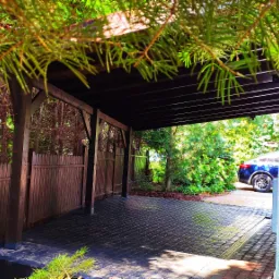 Zadaszone miejsce parkingowe z drewnianą konstrukcją, wyłożone kostką brukową, otoczone zielenią i drewnianym ogrodzeniem, z widocznym fragmentem zaparkowanego samochodu.