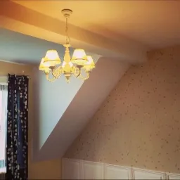 Pokój na poddaszu z beżową tapetą w gwiazdki, białym żyrandolem z abażurami, oknem z firankami sznurkowymi i ciemną zasłoną w kwiaty.