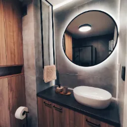 Nowoczesna łazienka z owalnym lustrem podświetlanym LED, umywalką nablatową i drewnianymi szafkami z czarnymi uchwytami, widoczny fragment kabiny prysznicowej w tle.