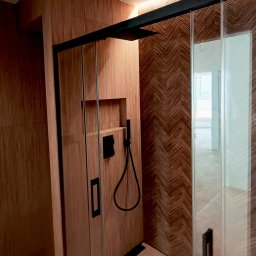 REMONT MIKO Wojciech Mikołajczyk - Nowoczesna kabina prysznicowa z drewnopodobnymi płytkami ułożonymi w jodełkę na ścianie, czarne detale armatury i oświetlenie LED. Minimalistyczny design.