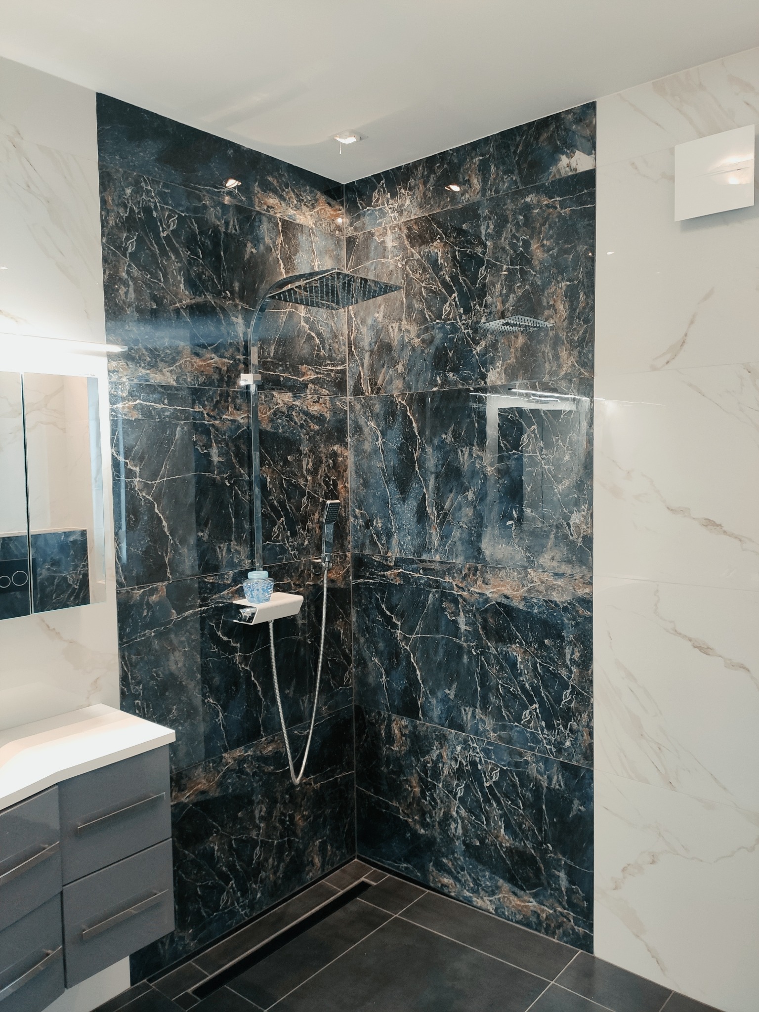 Nowoczesna kabina prysznicowa z deszczownicą, wyłożona płytami imitującymi czarny marmur z wyraźnymi żyłami, kontrastująca z jasnymi płytami na sąsiedniej ścianie, szara szafka umywalkowa...