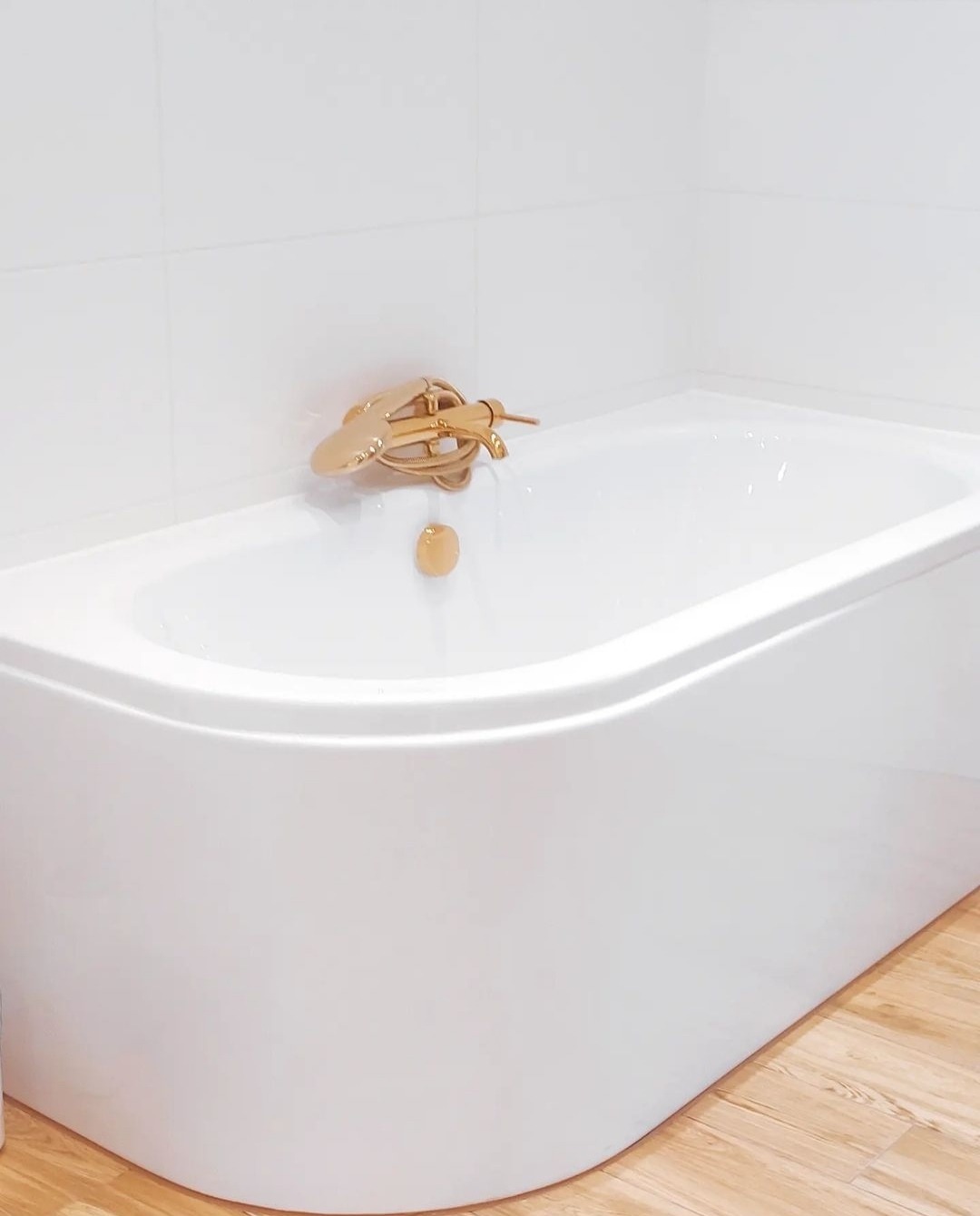 Biała wanna z zaokrąglonym frontem i złotą baterią w nowoczesnej łazience wykończonej białymi płytkami i drewnopodobną podłogą.