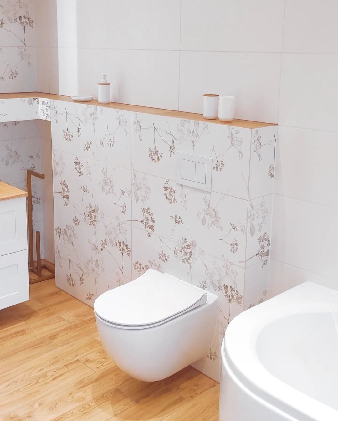 Nowoczesna łazienka z podwieszaną toaletą i płytkami w kwiatowy wzór, drewniane akcenty blatu i podłogi, minimalistyczne dodatki.