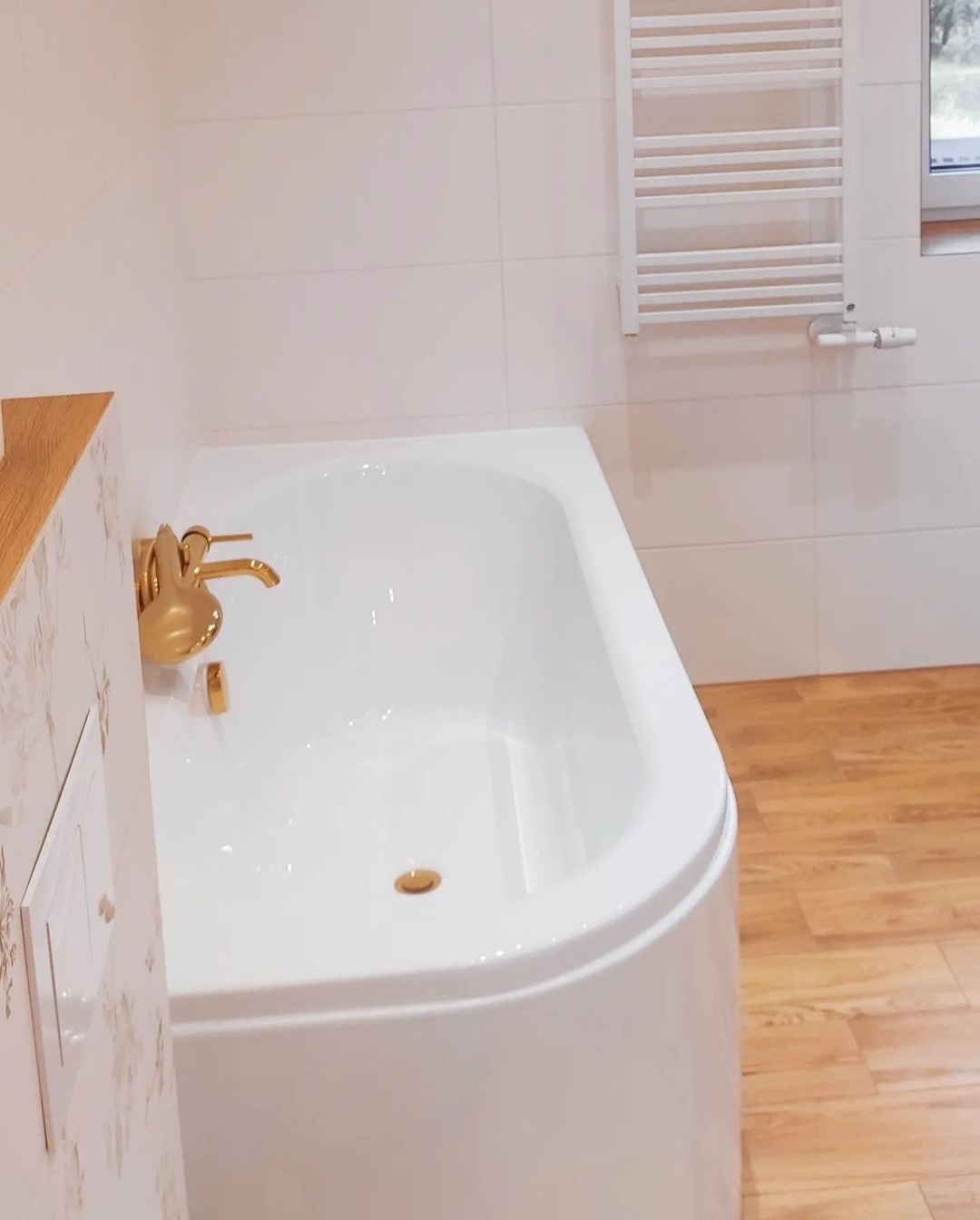 Biała wanna z narożną armaturą w kolorze złotym, jasne płytki na ścianach i drewnopodobna podłoga, grzejnik drabinkowy w tle.