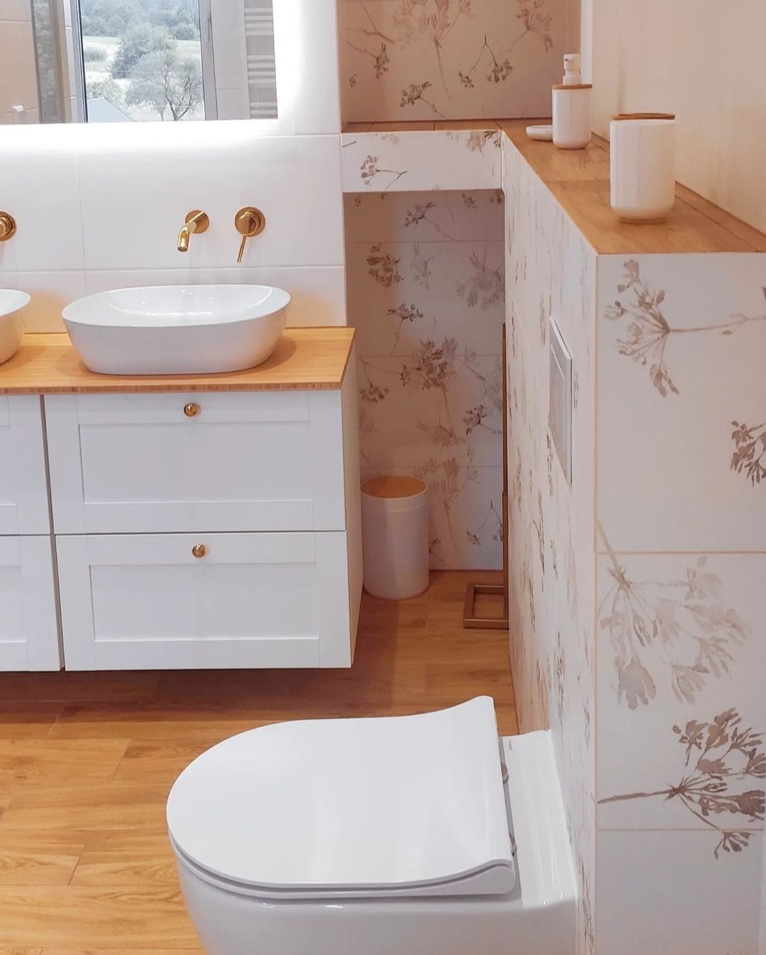Jasna łazienka z drewnianą podłogą, umywalką nablatową na szafce z białymi frontami, złotą armaturą i toaletą typu Rimless, wykończona płytkami w kwiatowe wzory.