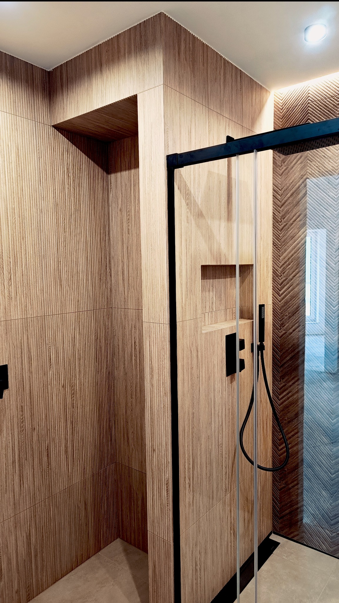 Nowoczesna kabina prysznicowa z drewnopodobnymi płytkami w odcieniach beżu, czarną armaturą i szklanymi drzwiami. Widoczne detale wykończenia i oświetlenie.