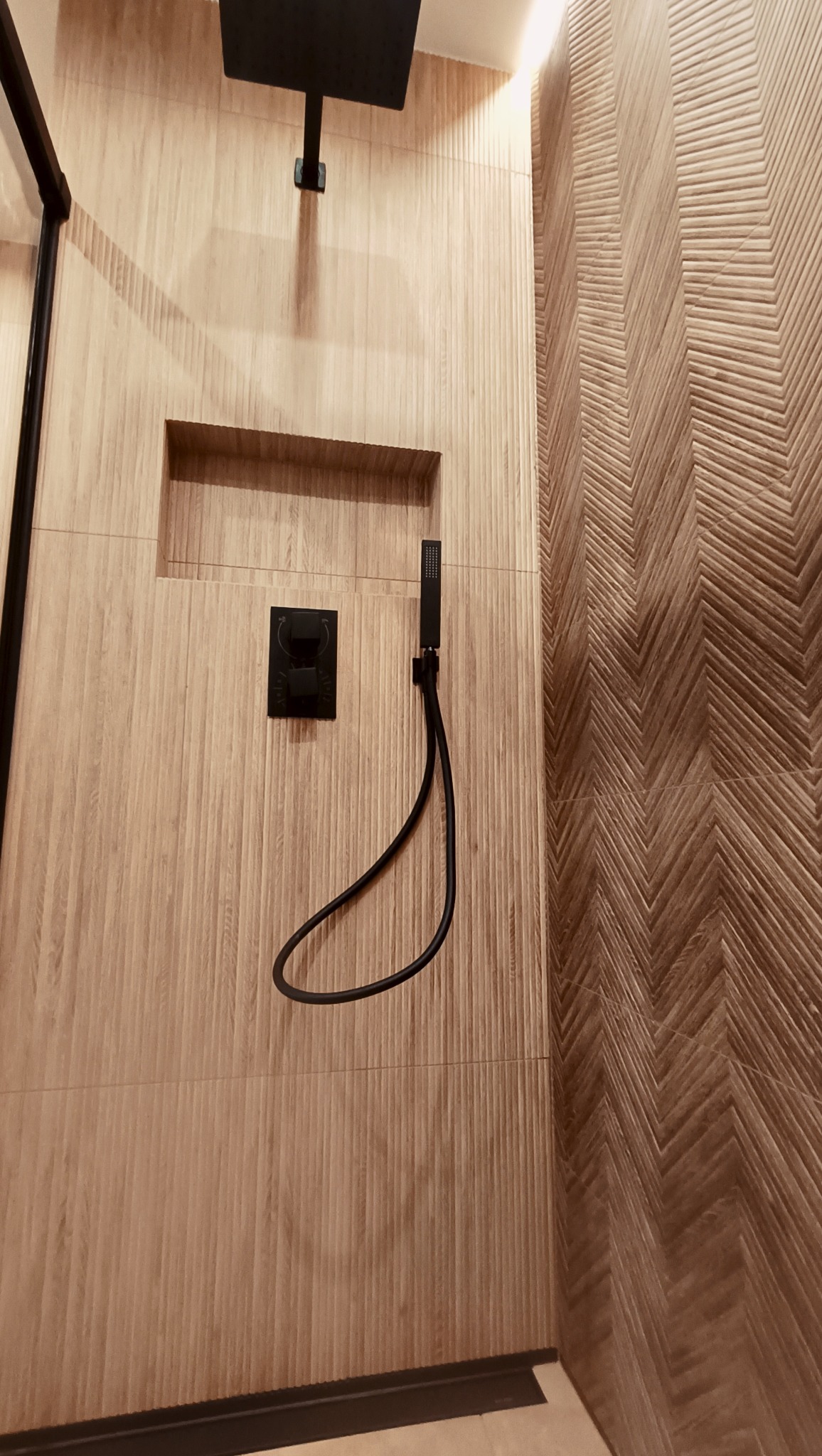 Nowoczesna łazienka z beżowymi płytkami imitującymi drewno, czarną armaturą i deszczownicą. Ściana z wzorem jodełki. Wnęka w ścianie.