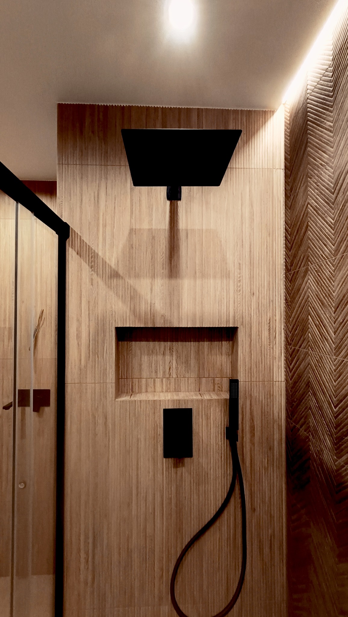 Nowoczesna łazienka z drewnopodobnymi płytkami, czarną armaturą i deszczownicą. Dekoracyjna ściana z wzorem jodełki. Oświetlenie LED podkreśla ciepły klimat.