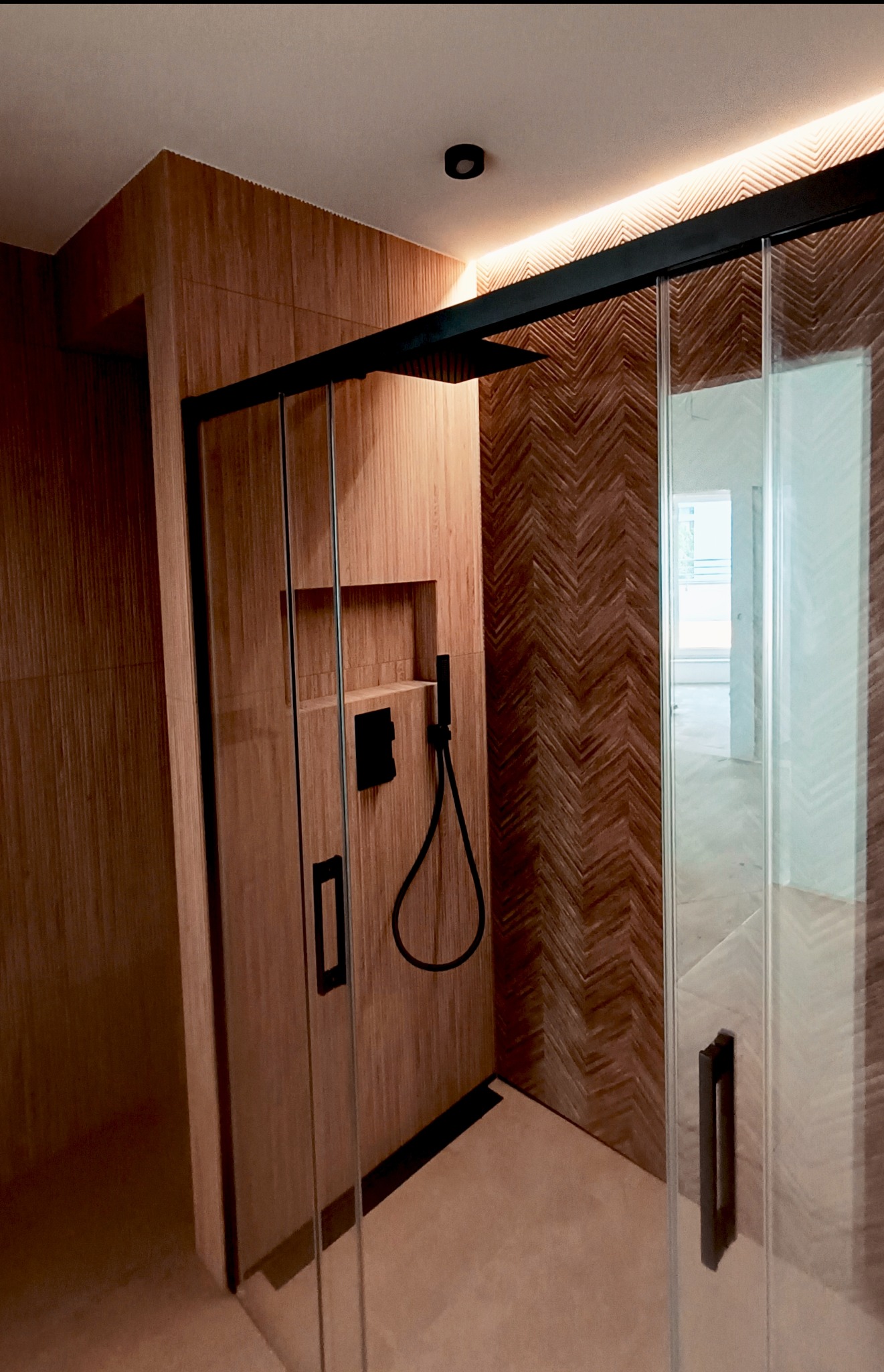 Nowoczesna kabina prysznicowa z drewnopodobnymi płytkami ułożonymi w jodełkę na ścianie, czarne detale armatury i oświetlenie LED. Minimalistyczny design.