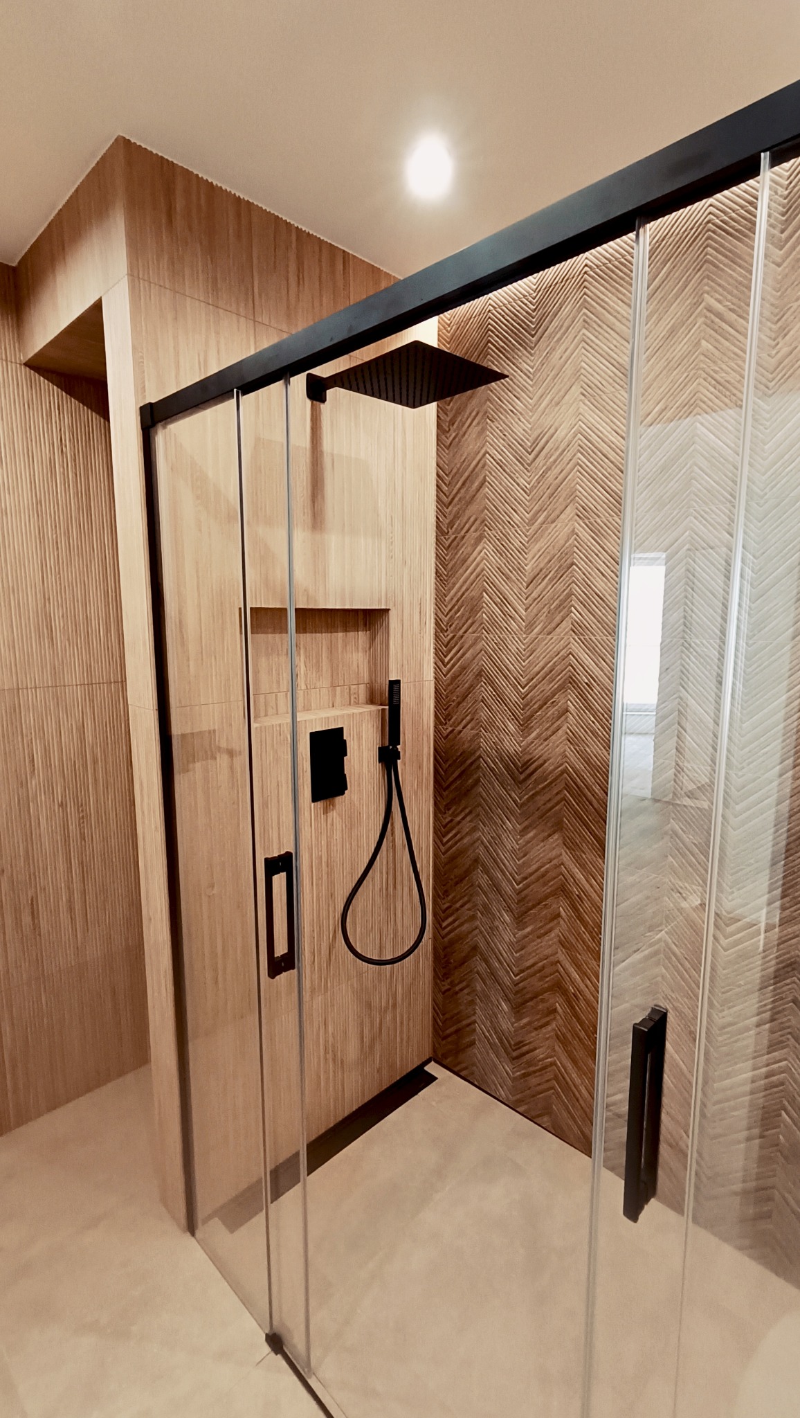 Nowoczesna kabina prysznicowa z drewnopodobnymi płytkami ułożonymi w jodełkę na ścianach. Czarna armatura i minimalistyczne uchwyty dodają elegancji.