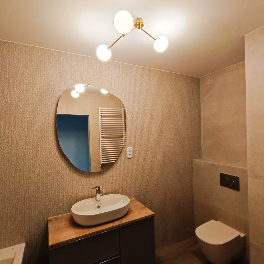 Wykończona łazienka z owalnym lustrem, umywalką na drewnianym blacie i nowoczesną lampą sufitową. Beżowe płytki na ścianach i podwieszana toaleta.
