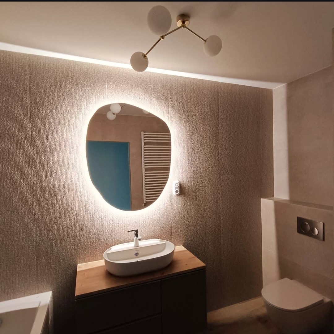 Nowoczesna łazienka z asymetrycznym lustrem podświetlanym LED, umywalką nablatową i minimalistyczną armaturą. Beżowe płytki i oświetlenie punktowe.