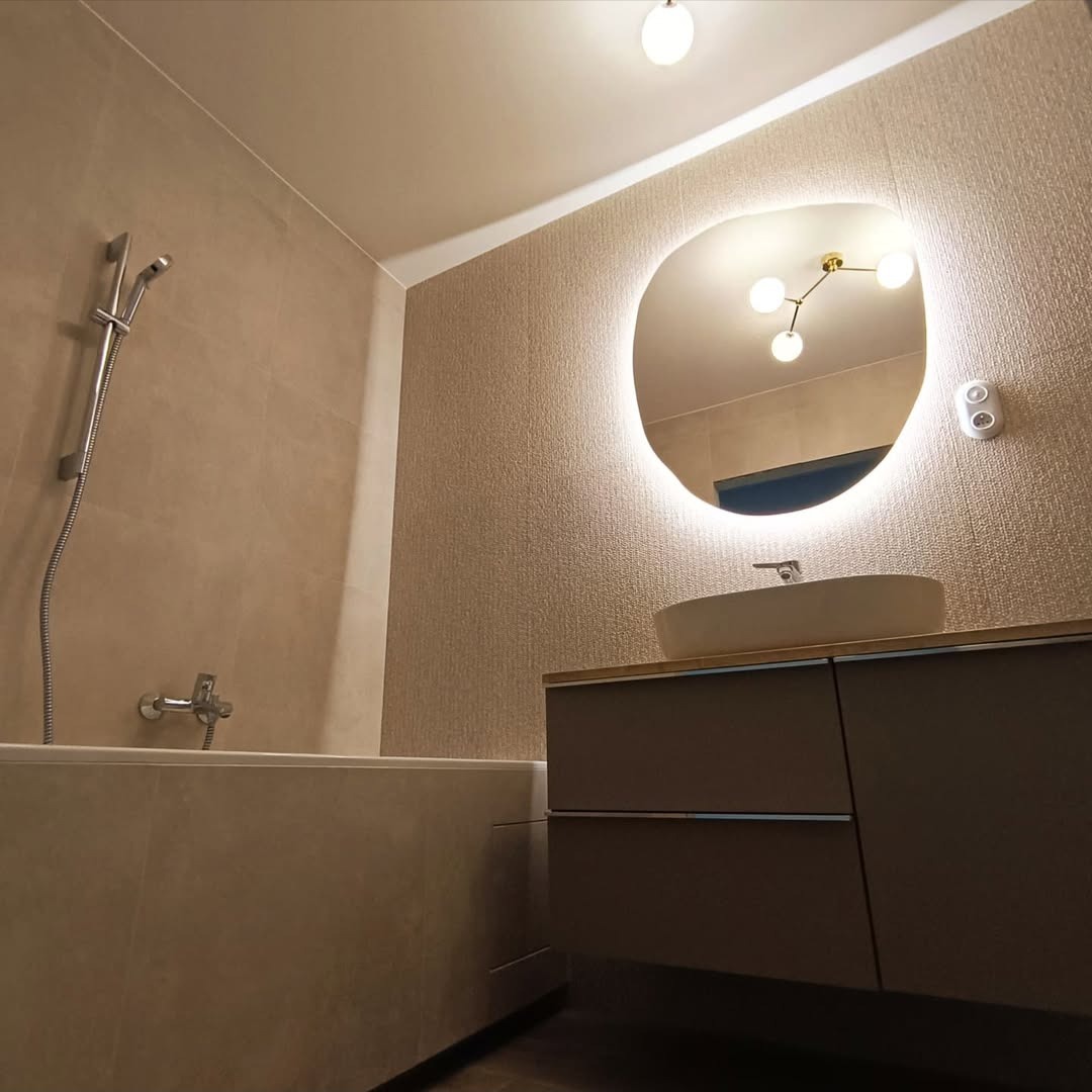 Wykończona łazienka w beżowych tonacjach z podświetlanym lustrem o nieregularnym kształcie, umywalką nablatową i zabudową meblową.