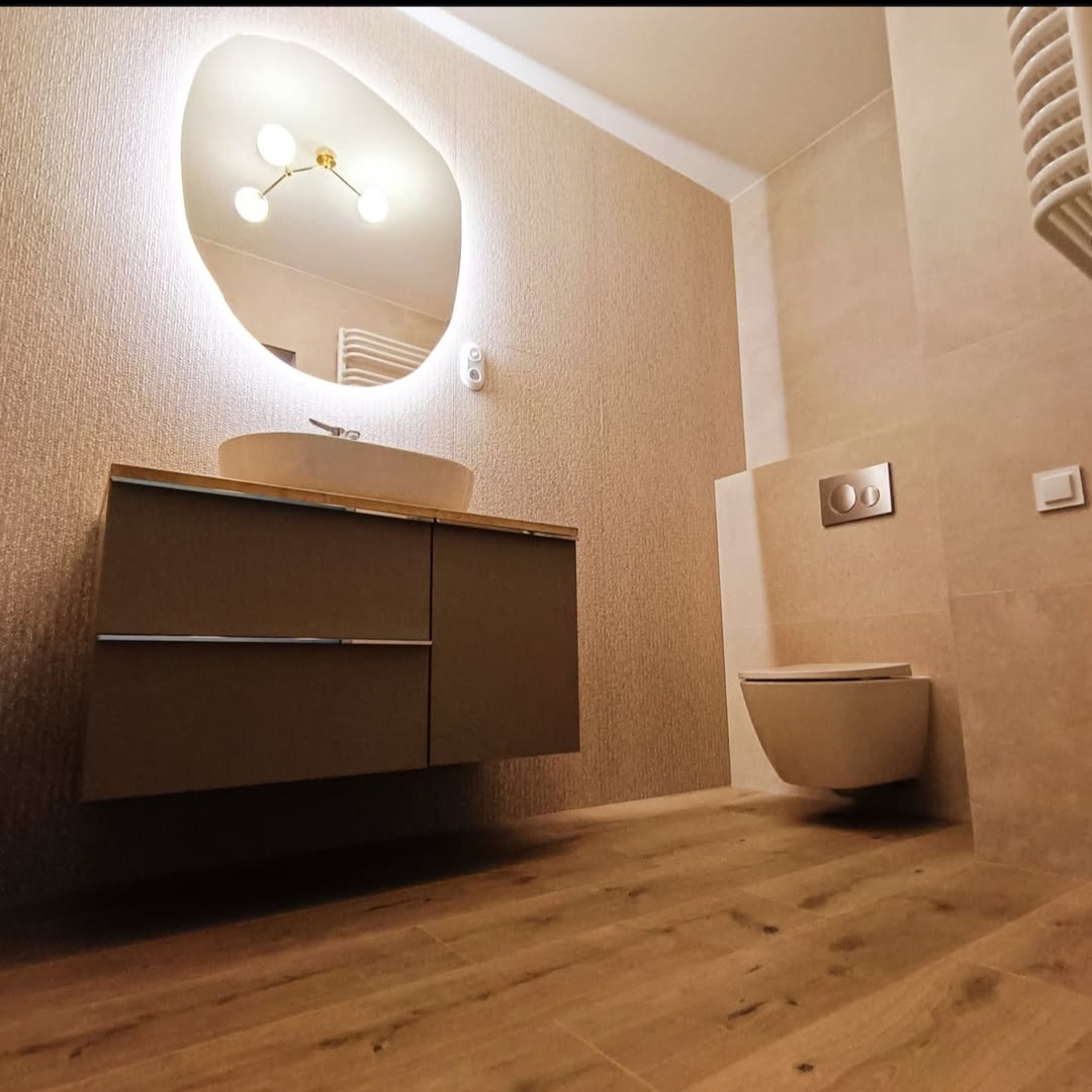 Nowoczesna łazienka z podwieszaną toaletą, umywalką na szafce i lustrem z podświetleniem LED. Beżowe płytki i drewnopodobna podłoga tworzą spójną całość.