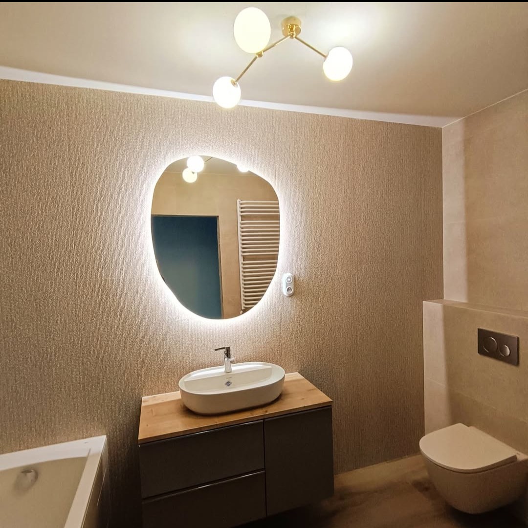 Nowoczesna łazienka po remoncie w Krakowie: asymetryczne lustro z podświetleniem LED, umywalka nablatowa na szafce z drewnianym blatem, beżowe płytki i designerska lampa.