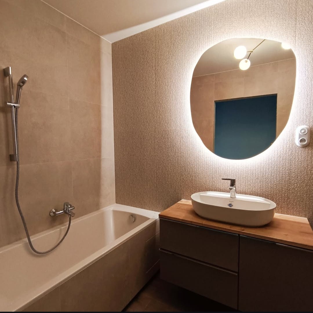 Nowoczesna łazienka z wanną, umywalką nablatową na szafce z drewnianym blatem i podświetlanym lustrem o nieregularnym kształcie. Beżowe płytki i teksturowana tapeta.