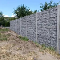 Długi, szary, betonowy płot z imitacją cegły oddzielający posesję z domem w tle od zaniedbanego terenu z ziemią i trawą.