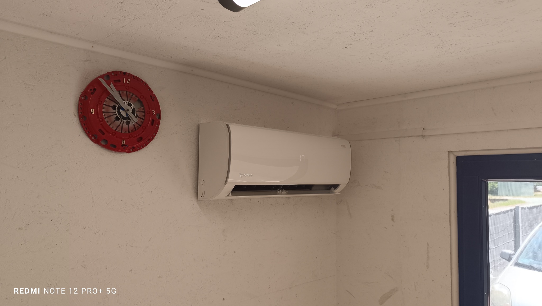 Biały klimatyzator Rotenso na ścianie z zegarem w kształcie sprzęgła BMW, w pomieszczeniu z oknem. Widoczny fragment samochodu za oknem.