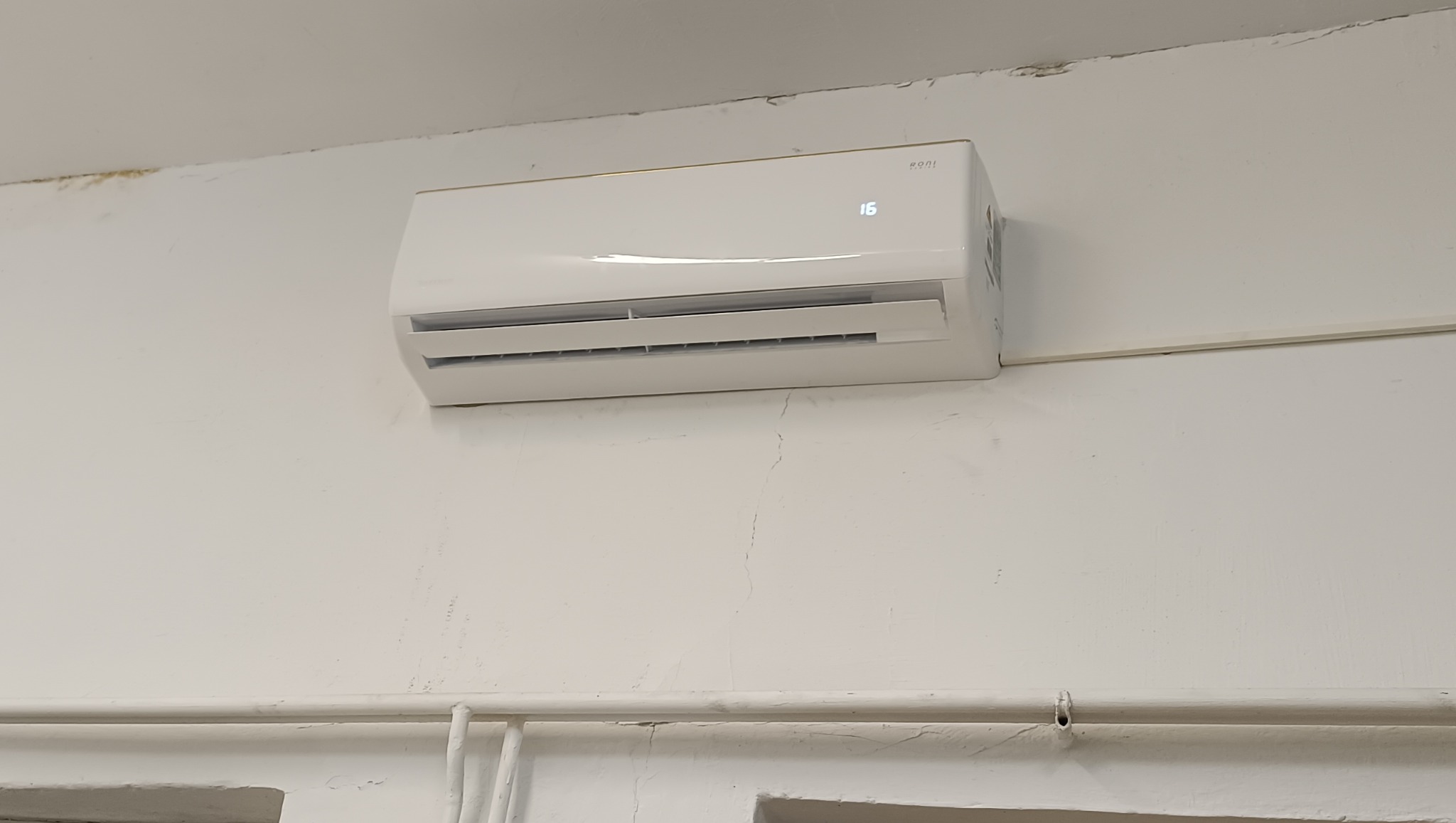 Biały klimatyzator ścienny marki Roni, ustawiony na 16 stopni, zamontowany na białej ścianie z widocznymi rurami instalacyjnymi w dolnej części kadru.