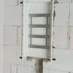 Biała, pusta skrzynka elektryczna z metalowymi szynami montażowymi, osadzona w surowej ścianie z betonu komórkowego z widocznymi ubytkami i resztkami zaprawy.