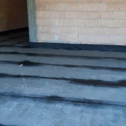 Izolacja przeciwwilgociowa fundamentów w trakcie realizacji, widoczne warstwy papy na betonowej wylewce oraz fragment ściany z pustaków ceramicznych.