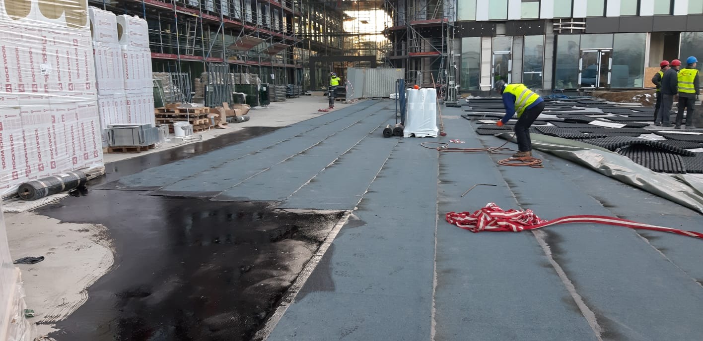 Prace izolacyjne fundamentów papą termozgrzewalną na dachu budynku w trakcie budowy, widoczne rolki materiału izolacyjnego, narzędzia i ekipa w odblaskowych kamizelkach.