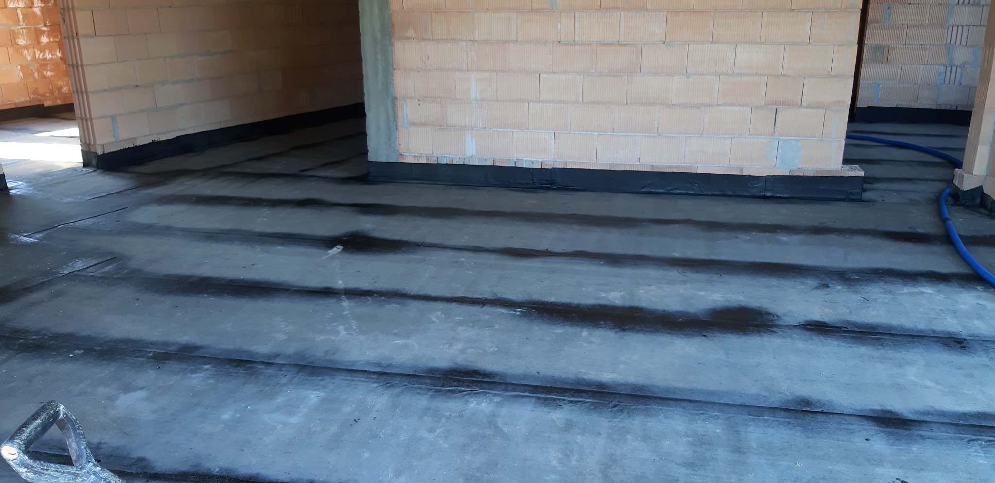 Izolacja przeciwwilgociowa fundamentów w trakcie realizacji, widoczne warstwy papy na betonowej wylewce oraz fragment ściany z pustaków ceramicznych.