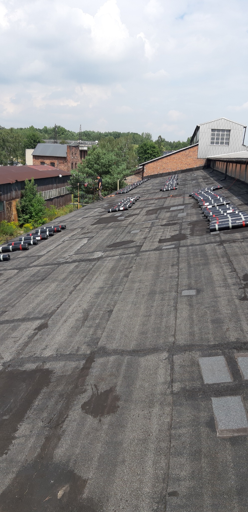Przygotowany do zgrzewania dach przemysłowy z rozłożonymi rolkami papy termozgrzewalnej; widoczny dekarz na rusztowaniu w oddali, otoczenie stanowią budynki przemysłowe i zieleń.