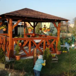 Drewniana altana z brązowym dachem, ozdobiona wiszącymi doniczkami z żółtymi kwiatami, widoczna zza siatki ogrodzeniowej w Górze, z małą postacią dziecka w tle.