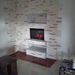 MOTO-LUK - Ściana wykończona dekoracyjnym kamieniem, z wbudowanym kominkiem elektrycznym z efektem płomieni, podłoga wyłożona płytkami imitującymi drewno, widoczny fragment mebla i stolika roboczego.