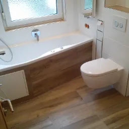 Nowoczesna łazienka z drewnopodobnymi płytkami na podłodze i obudowie wanny, biała armatura, podwieszana toaleta i okno z matową szybą, widoczna kratka odpływowa w podłodze.