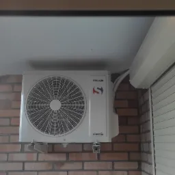 Biały klimatyzator Sinclair zamontowany na ceglanej ścianie balkonu, widoczne rury i roleta okienna.