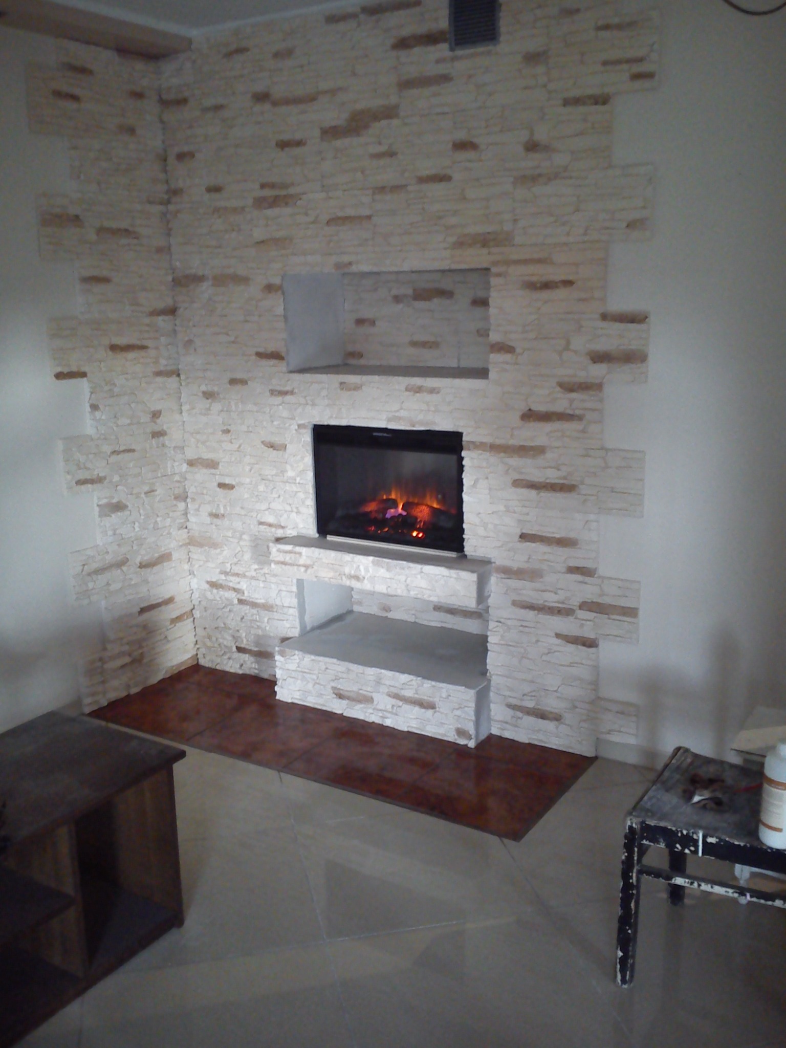 Ściana wykończona dekoracyjnym kamieniem, z wbudowanym kominkiem elektrycznym z efektem płomieni, podłoga wyłożona płytkami imitującymi drewno, widoczny fragment mebla i stolika roboczego.