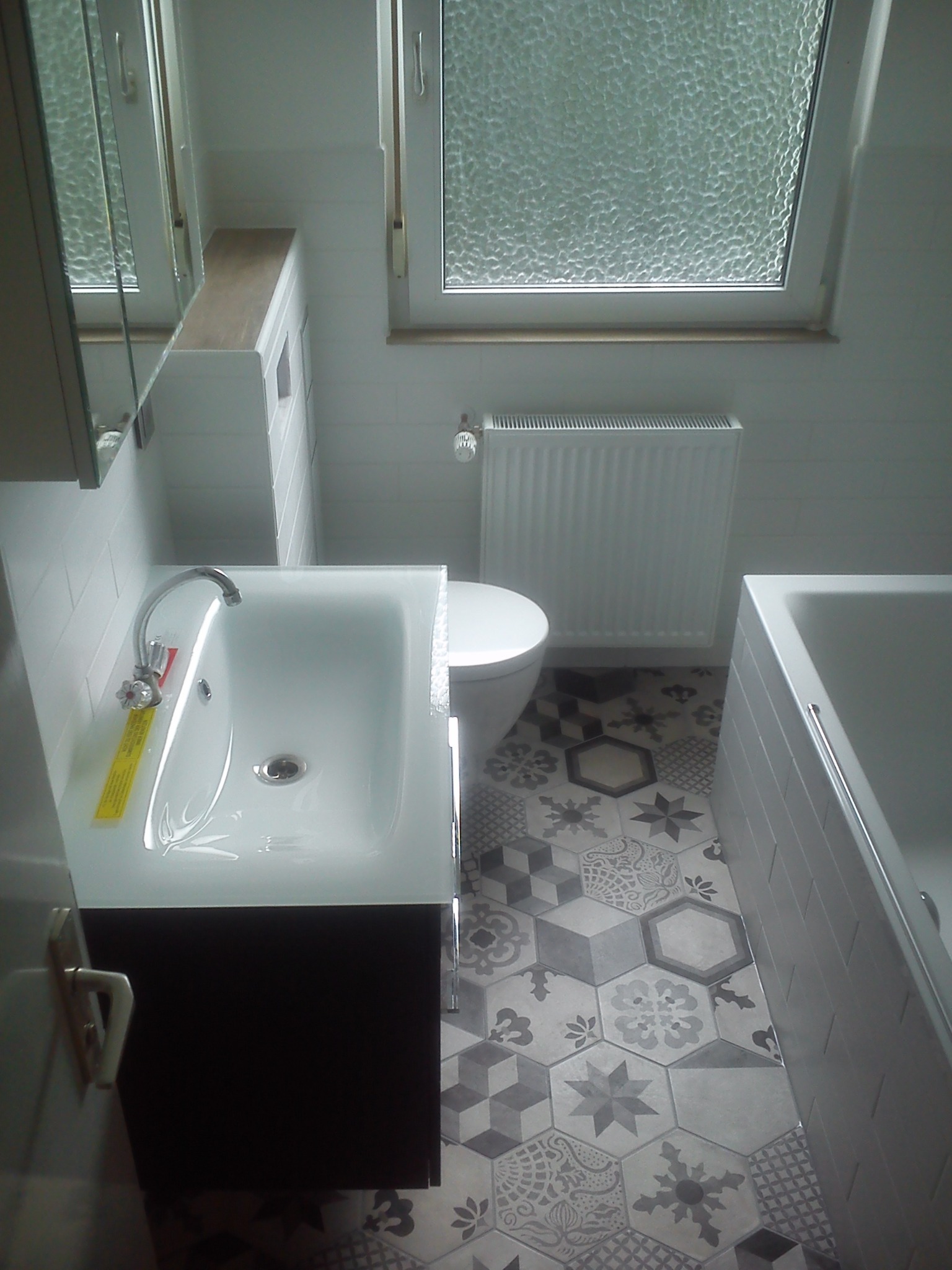 Umywalka z szafką w odnowionej łazience, armatura z żółtą etykietą, wzorzysta podłoga z płytek heksagonalnych, białe płytki na ścianach, okno z szybą ornamentową.