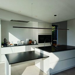 KR-Construct - Nowoczesna, jasna kuchnia z wyspą i minimalistycznym oświetleniem LED. Kontrast czarnego blatu i białych szafek. Uwagę przykuwa długa, pozioma lampa wisząca.