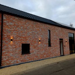 KR-Construct - Elewacja budynku z czerwonej cegły z czarnym dachem i oknami, oświetlona zewnętrznymi lampami ściennymi, z panelami słonecznymi na dachu. Widok z poziomu ulicy.