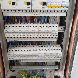 KR-Construct - Rozdzielnia elektryczna w trakcie montażu. Widoczne bezpieczniki, przewody w różnych kolorach i złącza. Biała obudowa, surowe wykończenie ściany.