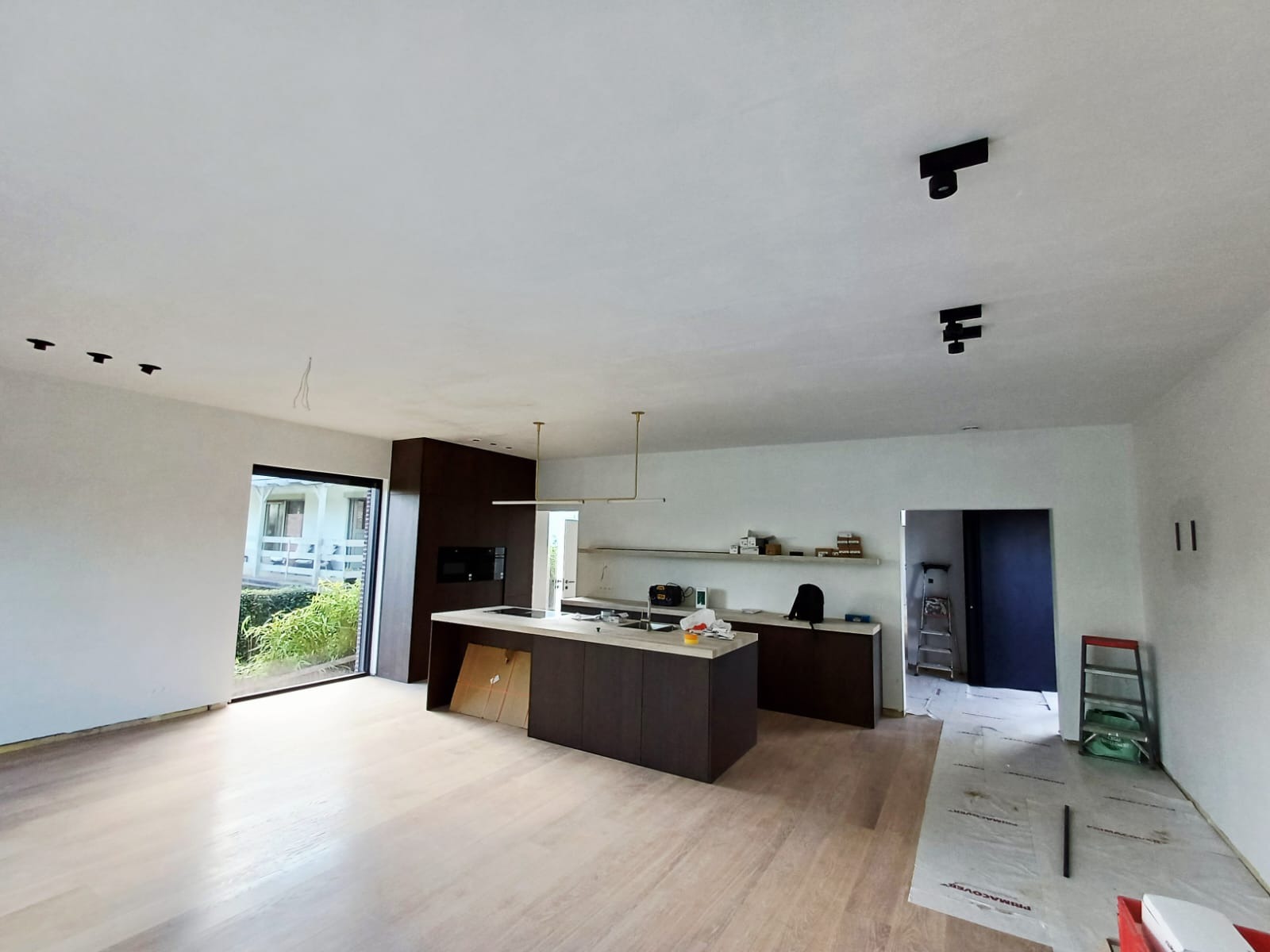 Nowoczesna kuchnia z wyspą i oświetleniem punktowym, widok na okno tarasowe i schody. Wnętrze w trakcie wykończenia, podłoga zabezpieczona folią.