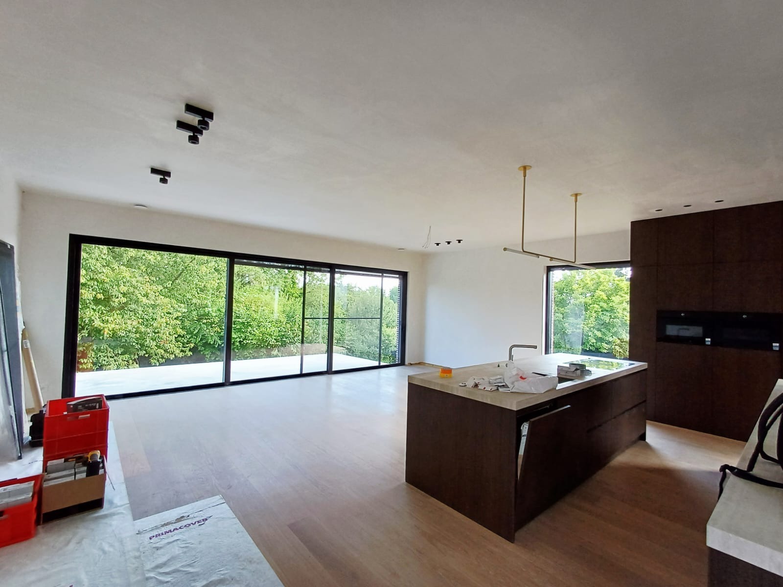 Przestronne wnętrze z dużymi oknami i wyspą kuchenną. Widoczne czarne lampy sufitowe i drewniana podłoga. W tle zieleń za oknem.