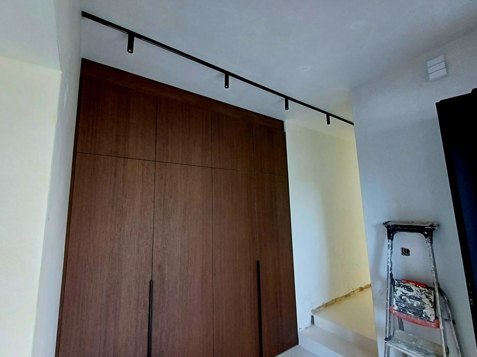 Wnętrze z garderobą z ciemnego drewna i czarnym systemem szynowym oświetlenia punktowego na suficie. Widoczna drabina malarska.