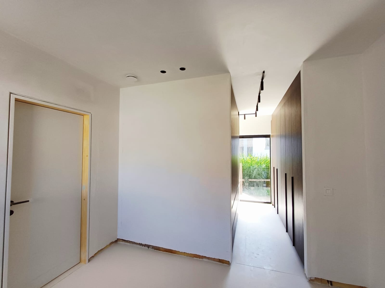 Wnętrze w trakcie wykańczania: białe ściany, surowa podłoga, widoczne punkty oświetleniowe w suficie i czarna listwa oświetleniowa, drzwi z ościeżnicą w trakcie montażu.