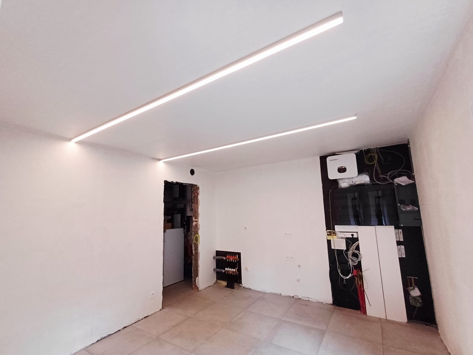 Nowoczesne oświetlenie liniowe LED na suficie w remontowanym pomieszczeniu z widoczną instalacją elektryczną i rozdzielnicą. Świeże, białe ściany i płytki podłogowe.