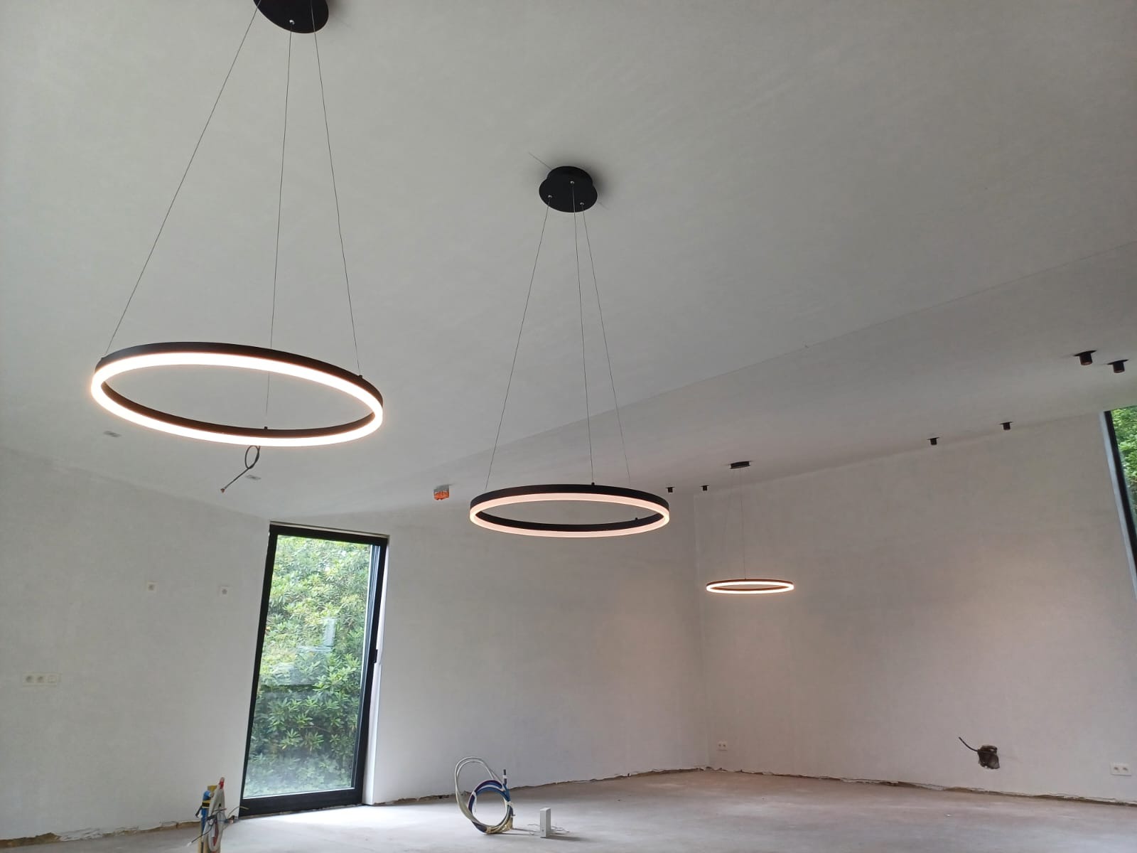 Nowoczesne, okrągłe lampy LED wiszące w surowym, jasnym wnętrzu. Minimalistyczny design oświetlenia podkreśla prostotę pomieszczenia z dużym oknem.