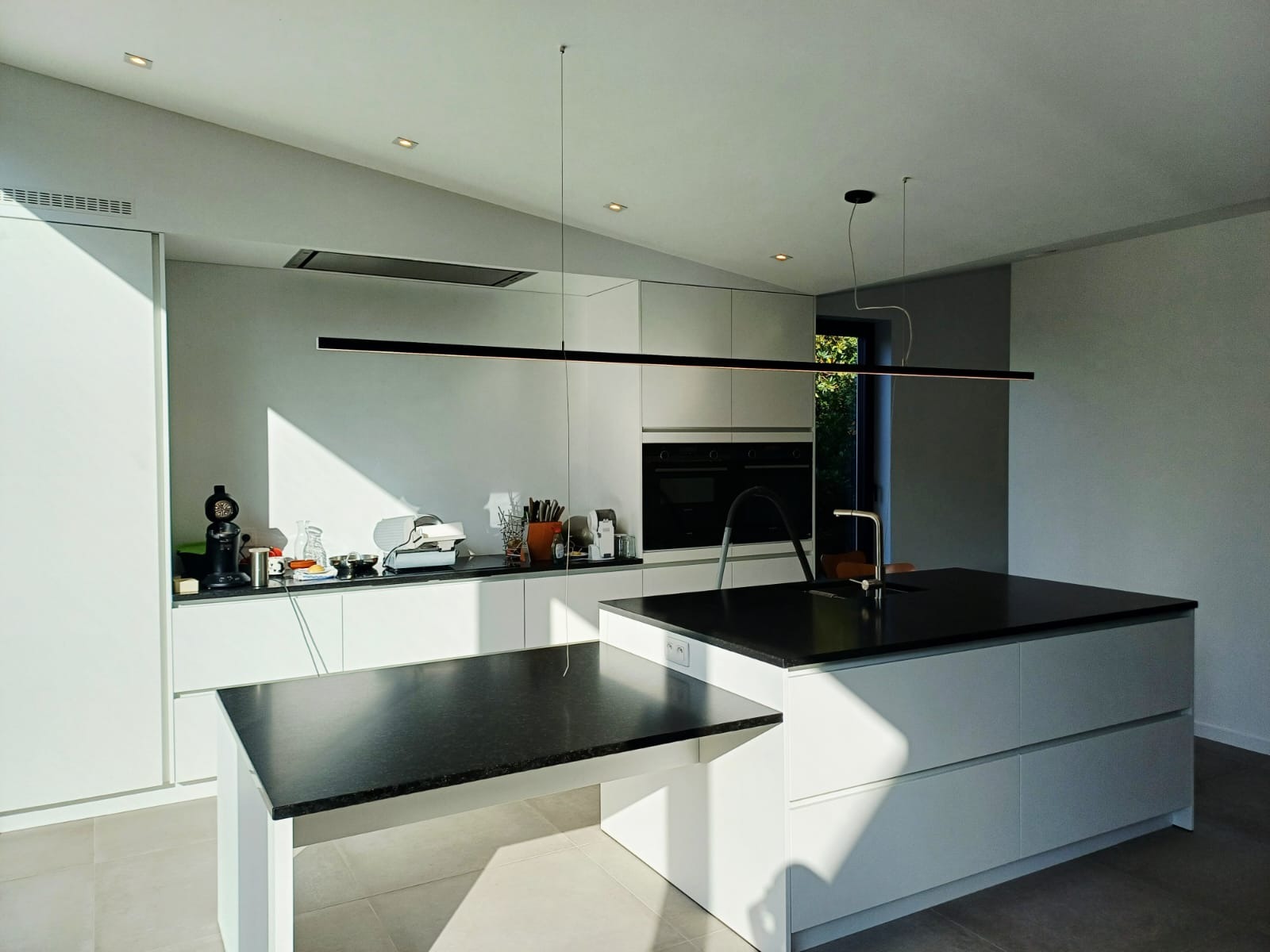 Nowoczesna, jasna kuchnia z wyspą i minimalistycznym oświetleniem LED. Kontrast czarnego blatu i białych szafek. Uwagę przykuwa długa, pozioma lampa wisząca.