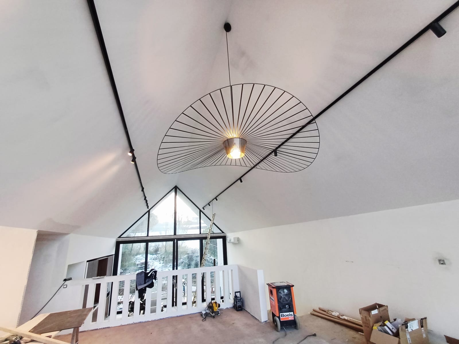 Nowoczesne oświetlenie sufitowe z dekoracyjną lampą wiszącą i szynoprzewodami w jasnym wnętrzu z dużym oknem trójkątnym i widocznymi narzędziami pracy.